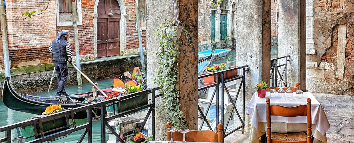 A gondolier rowing down a narrow canal in Venice, passing rustic buildings and a small outdoor dining table
